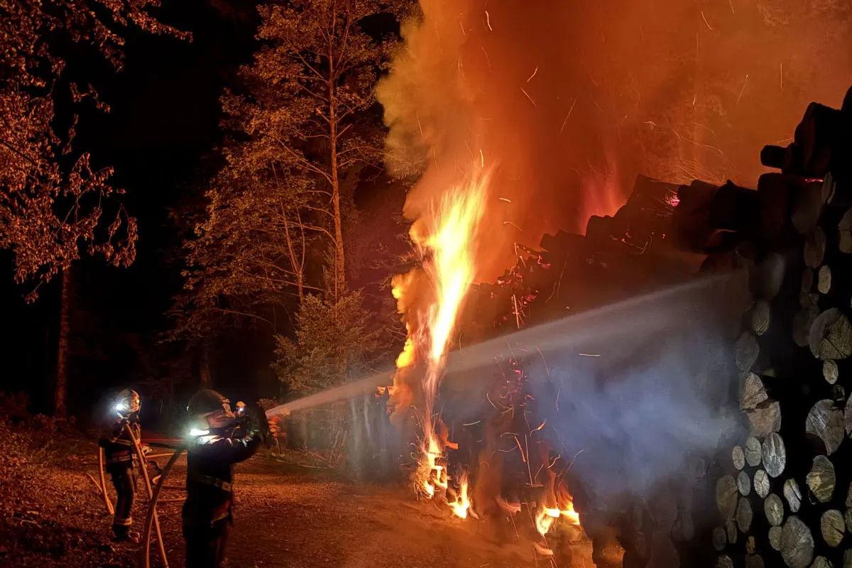 Zwei Feuerwehrleute stehen nachts im Freien und löschen einen großen brennenden Holzstapel mit einem Wasserstrahl.