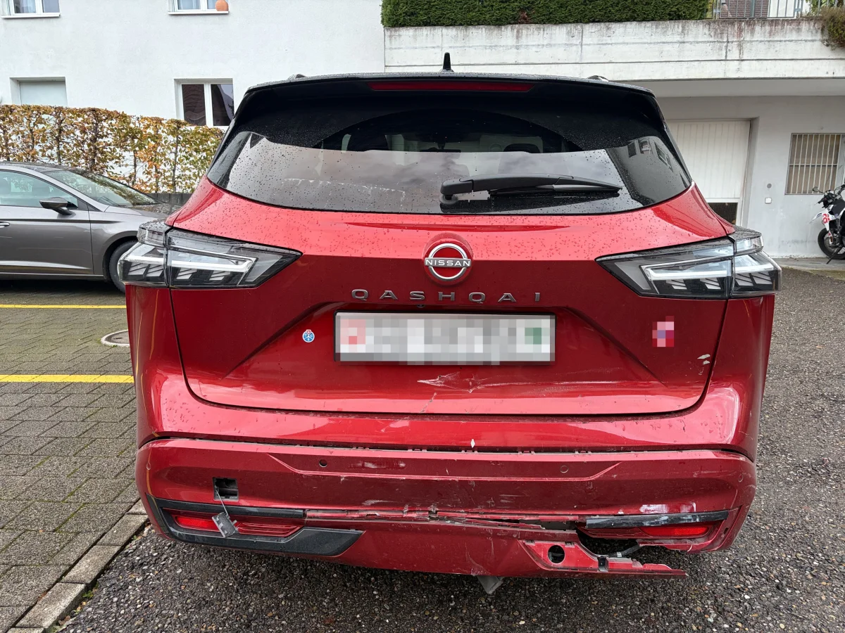 Ein roter Nissan Qashqai mit beschädigtem hinteren Stoßfänger steht auf einem Parkplatz. Im Hintergrund sind andere Fahrzeuge und eine Hauswand sichtbar.