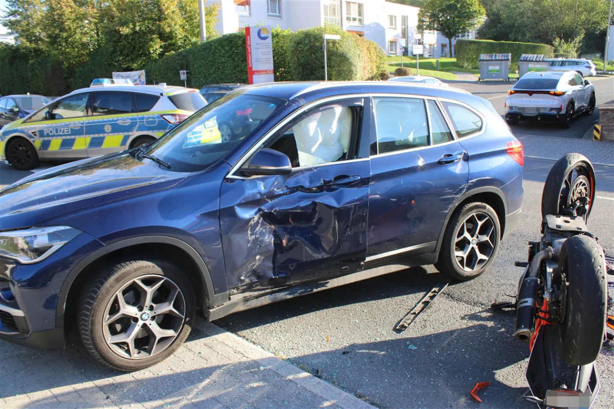 Ein blauer BMW mit zerbeulter Tür steht neben einem umgestürzten Motorrad auf der Straße. Im Hintergrund ist ein Polizeifahrzeug zu sehen.