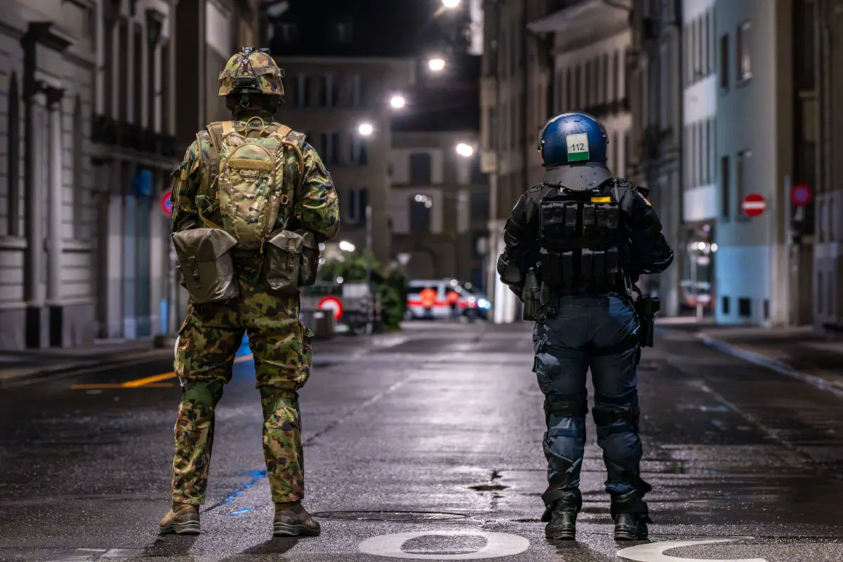 Zwei uniformierte Personen, eine in Tarnkleidung und eine in dunkler Uniform, stehen auf einer beleuchteten Straße bei Nacht.