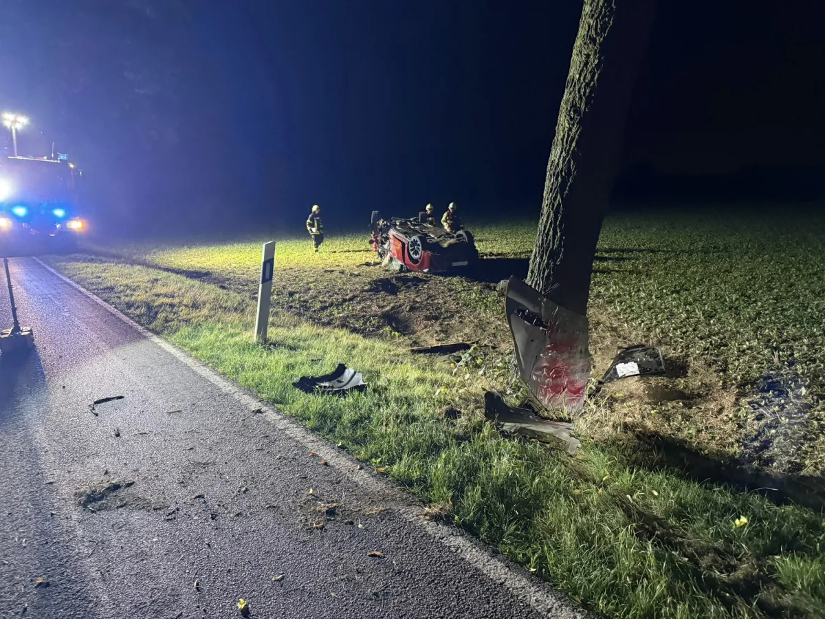 Das Bild zeigt ein rotes Auto, das nachts auf einer Wiese neben einem Baum auf dem Dach liegt. Feuerwehrleute sind im Einsatz, und ein Löschfahrzeug leuchtet die Szene von der Straße aus.