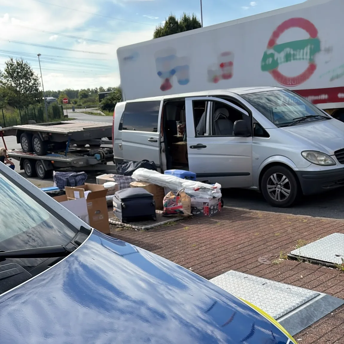 Ein silberner Van steht auf einem Parkplatz mit geöffneter Seitentür. Mehrere Gepäckstücke und Kartons sind daneben auf dem Boden verteilt.
