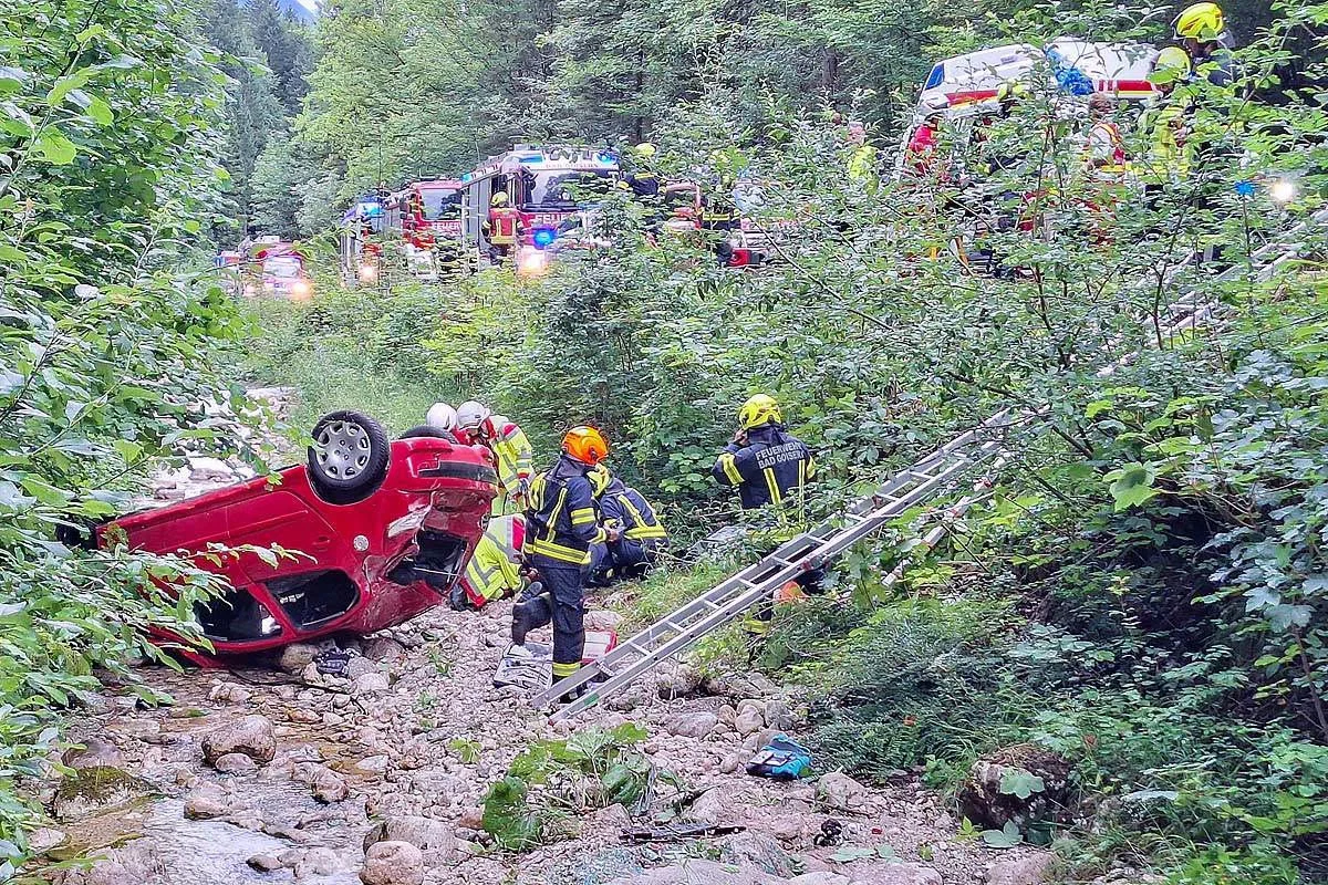 Ein rotes Auto liegt umgestürzt auf dem Dach in einem Bachbett, umgeben von Feuerwehrleuten in einem Waldgebiet.