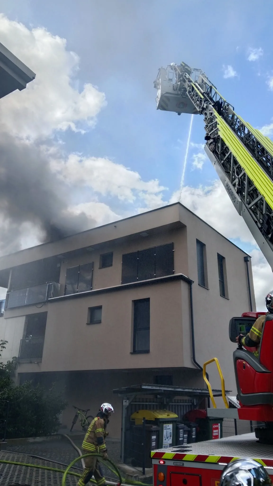Ein Feuerwehrmann löscht einen Brand in einem Wohngebäude. Eine Drehleiter spritzt Wasser über das Dach; Rauch steigt auf.