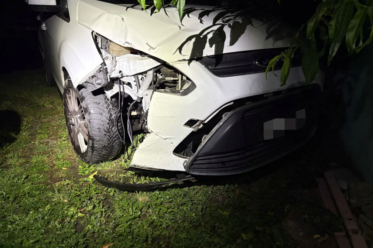 Ein weißes Auto mit schweren Frontschäden steht auf einer bewachsenen Fläche. Die Szene ist bei nächtlicher Beleuchtung aufgenommen.