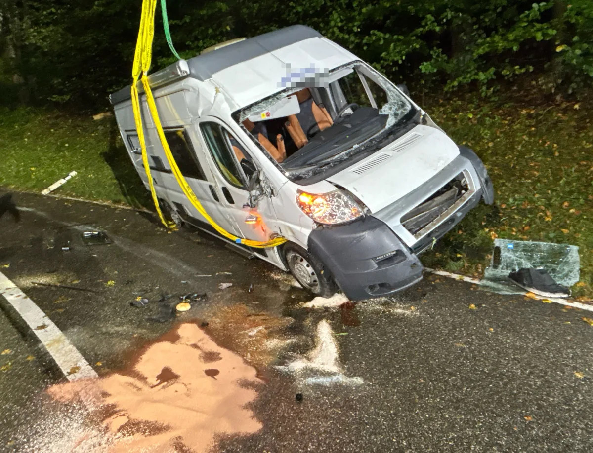 Ein verunfallter Lieferwagen liegt seitlich an einer Straßenböschung. Der Wagen ist beschädigt, und Trümmer liegen auf der Straße.