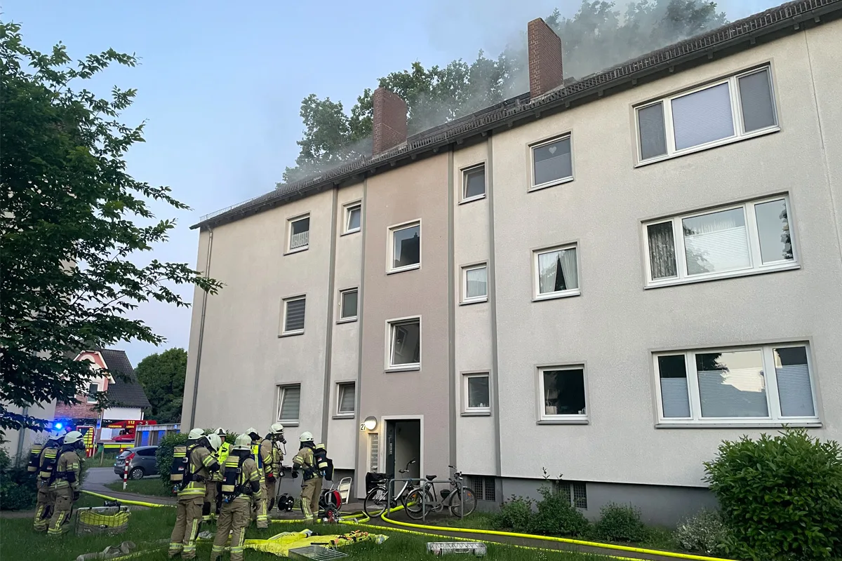Mehrstöckiges Wohngebäude, aus dessen Dach Rauch aufsteigt, während Feuerwehrleute in Einsatzkleidung davor stehen.