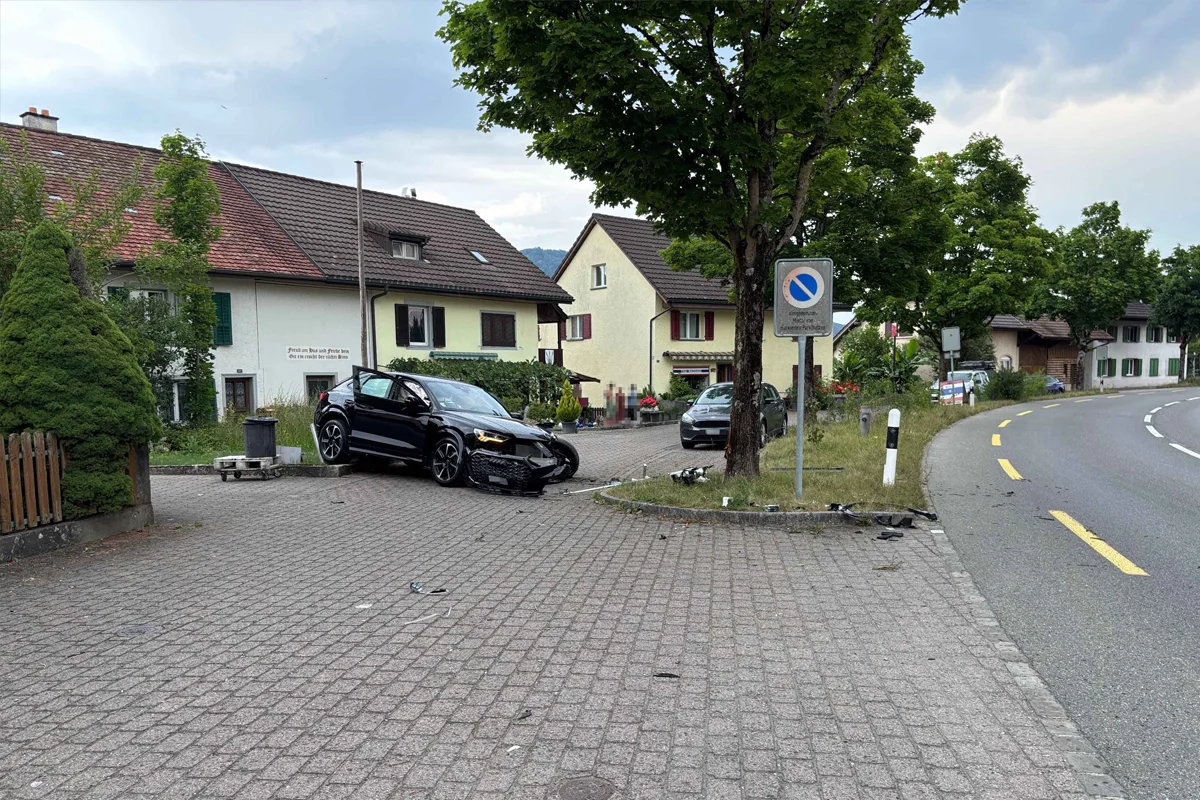 Ein schwarzes Auto mit erheblichen Frontschäden steht auf einem Gehweg in einer ländlichen Wohngegend. Im Hintergrund sind mehrere Wohnhäuser sichtbar.