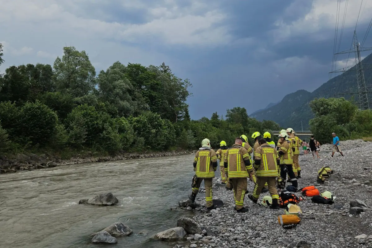 Eine Gruppe von Feuerwehrleuten in Einsatzkleidung befindet sich an einem Flussufer. Der Himmel ist bewölkt und deutet auf ein bevorstehendes Gewitter hin. Im Hintergrund sind bewaldete Berge sichtbar.
