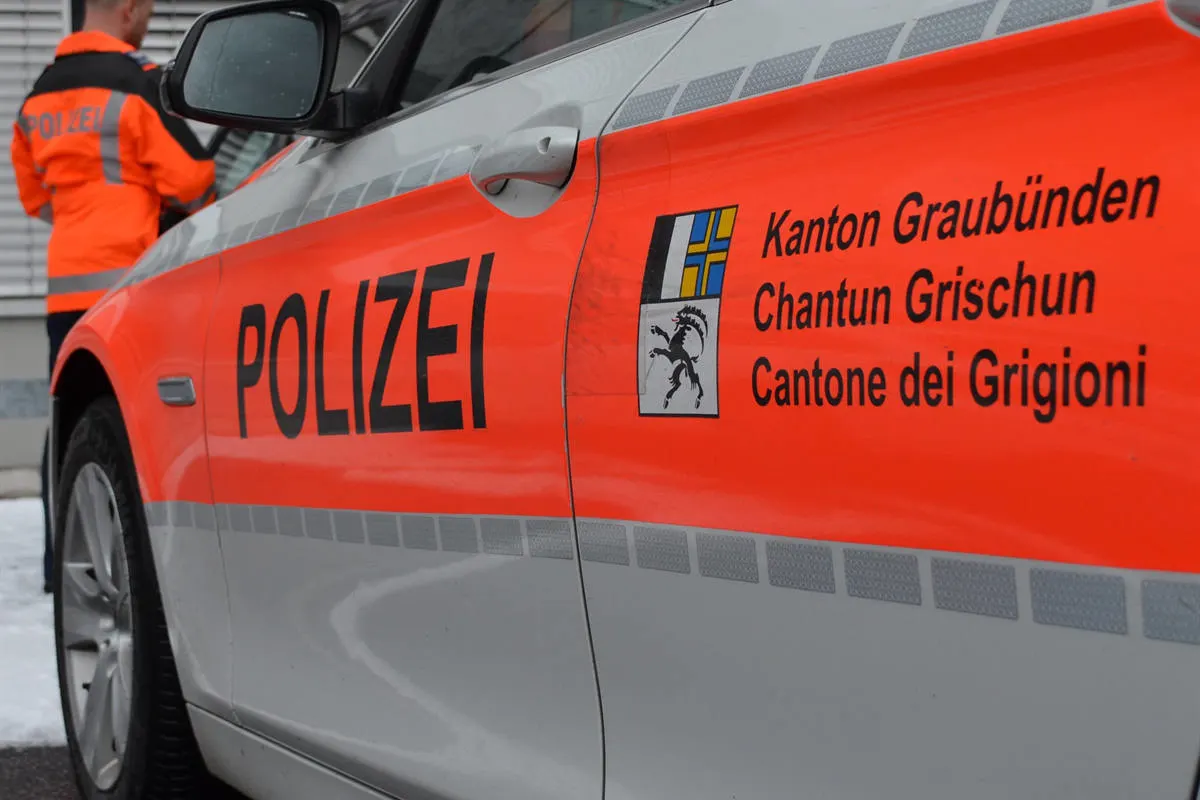 Ein rot-weißes Polizeifahrzeug der Kantonspolizei Graubünden mit sichtbarem Wappen steht im Schnee. Ein Polizist ist im Hintergrund erkennbar.