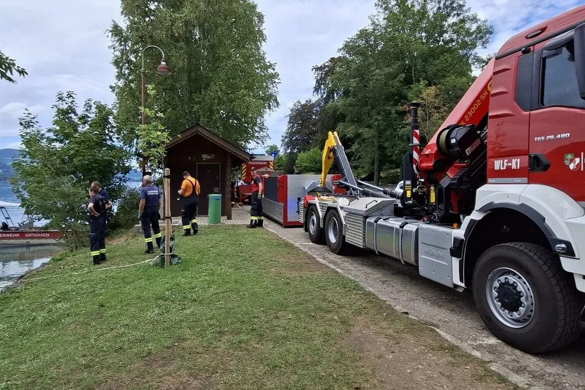 Mehrere Feuerwehrleute stehen neben einem großen Feuerwehr-Lkw am Ufer eines Sees. Ein kleiner Schuppen ist ebenfalls sichtbar.