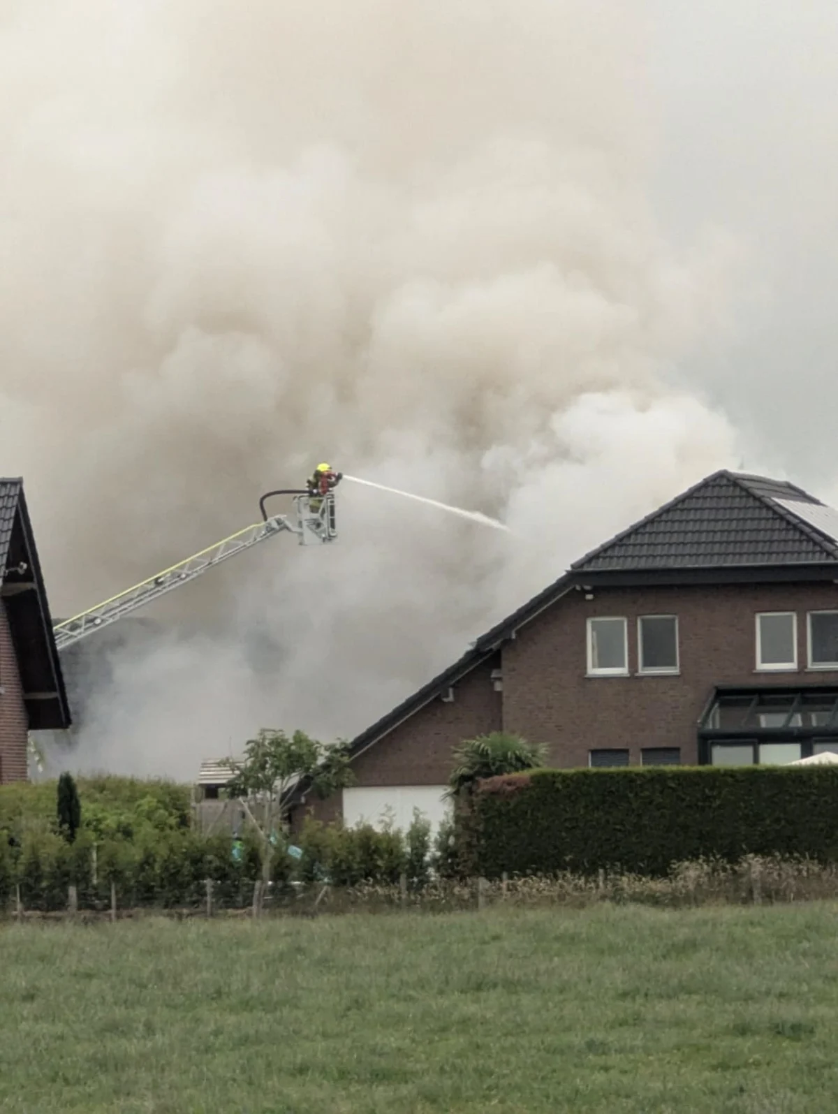 Ein Feuerwehrmann auf einer Leiter bekämpft einen Dachstuhlbrand mit einem Wasserstrahl, während Rauch aufsteigt.