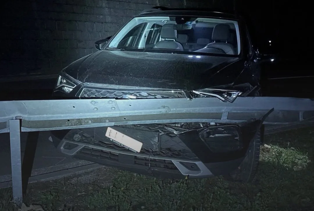 Ein dunkles Auto ist frontal gegen eine Leitplanke gefahren. Die Szene ist bei Nacht aufgenommen und zeigt deutliche Schäden am Fahrzeug.