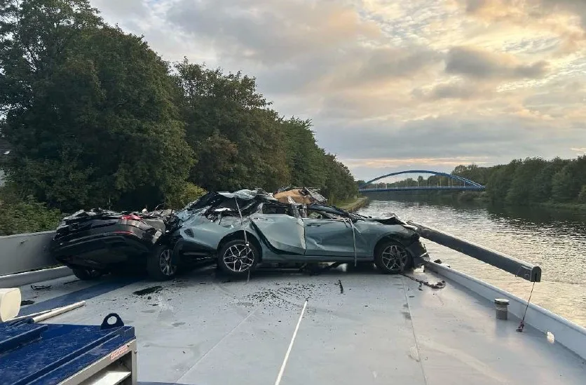Zwei stark beschädigte Autos stehen auf dem Deck eines Frachtschiffs. Im Hintergrund ist ein Fluss und eine Brücke zu sehen.