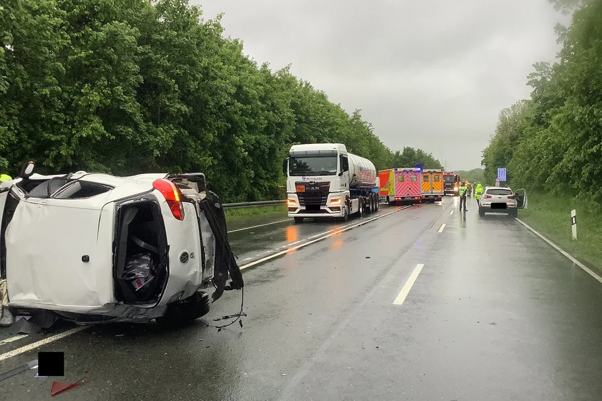 Ein weißer PKW liegt umgekippt auf der regennassen Autobahn. Im Hintergrund sind ein LKW und ein Rettungsfahrzeug zu sehen. Mehrere Personen in gelben Jacken stehen auf der Straße.
