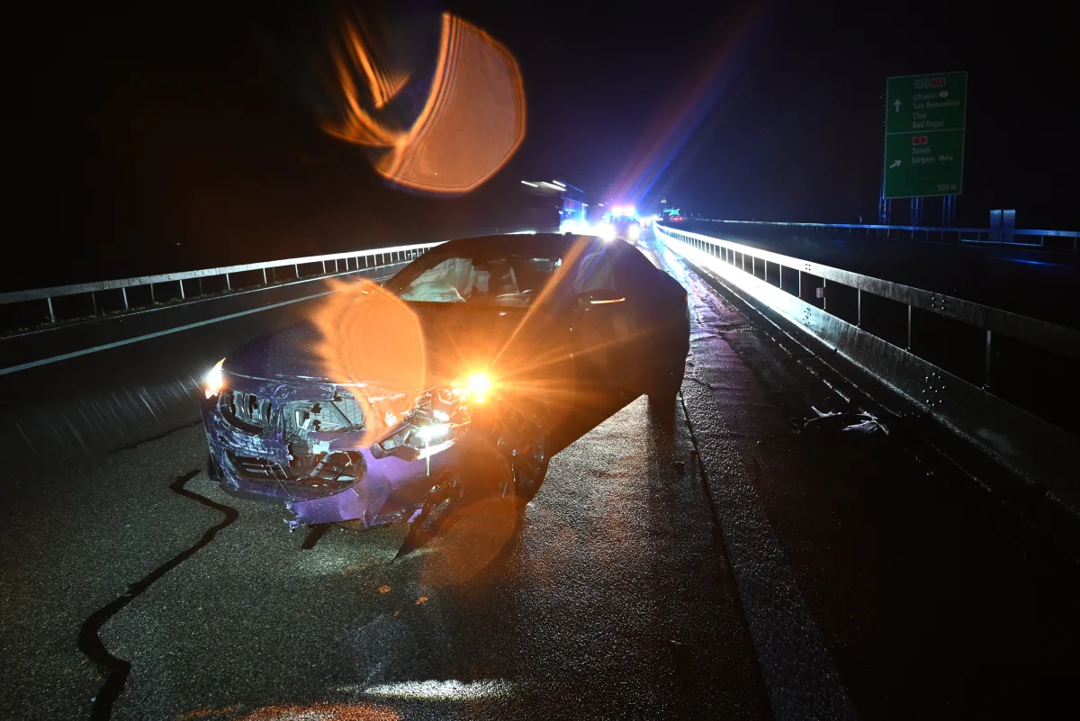 Ein Auto mit Frontschaden steht in der Dunkelheit auf einer nassen Autobahn, umgeben von reflektierenden Lichtern und Fahrzeugen im Hintergrund.