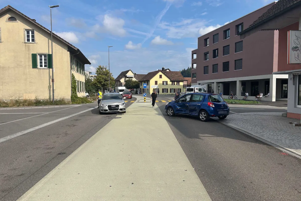 Ein Audi steht am Straßenrand, während ein beschädigter blauer Kleinwagen auf der Straße sichtbar ist. Menschen stehen in der Nähe der Fahrzeuge.