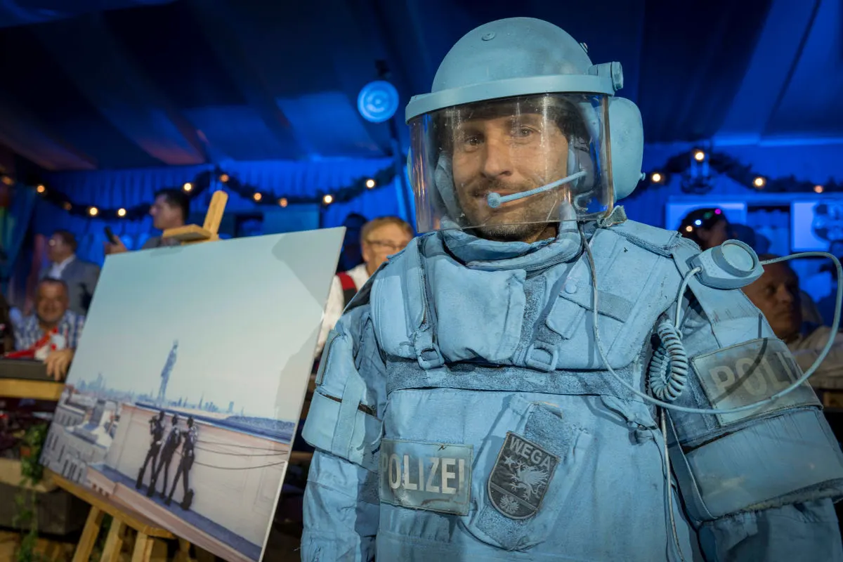 Ein Mann trägt eine blaue Schutzrüstung mit dem Schriftzug 'Polizei' und einem Wappen 'WEGA' in einem festlichen Umfeld.