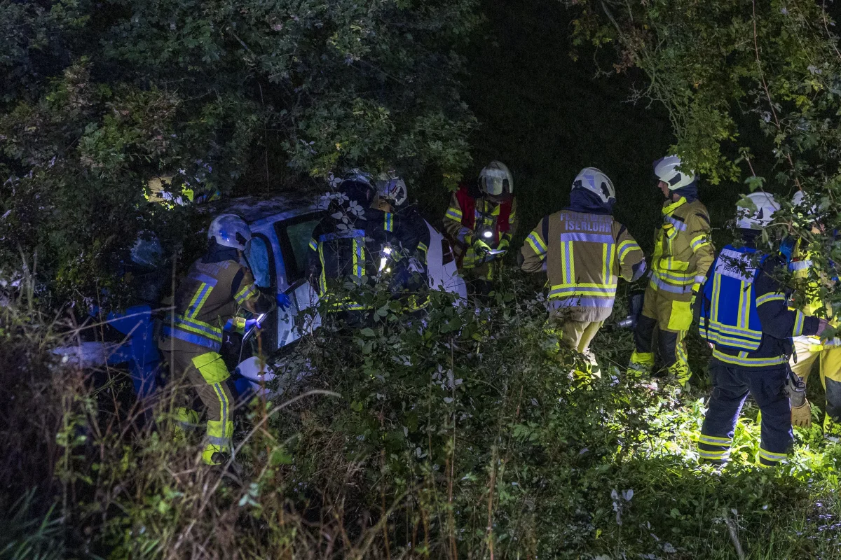 Mehrere Feuerwehrleute in gelber Schutzkleidung und Helmen arbeiten nachts an einem verunfallten Fahrzeug, das in dichtem Gebüsch steckt.