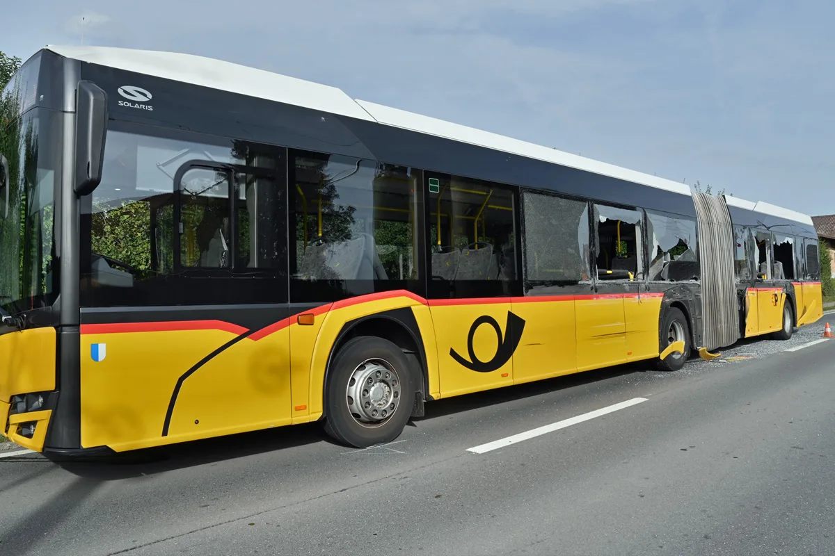 Ein gelber Postbus mit rot-schwarzen Streifen steht am Straßenrand. Die Fenster des Busses sind deutlich beschädigt.