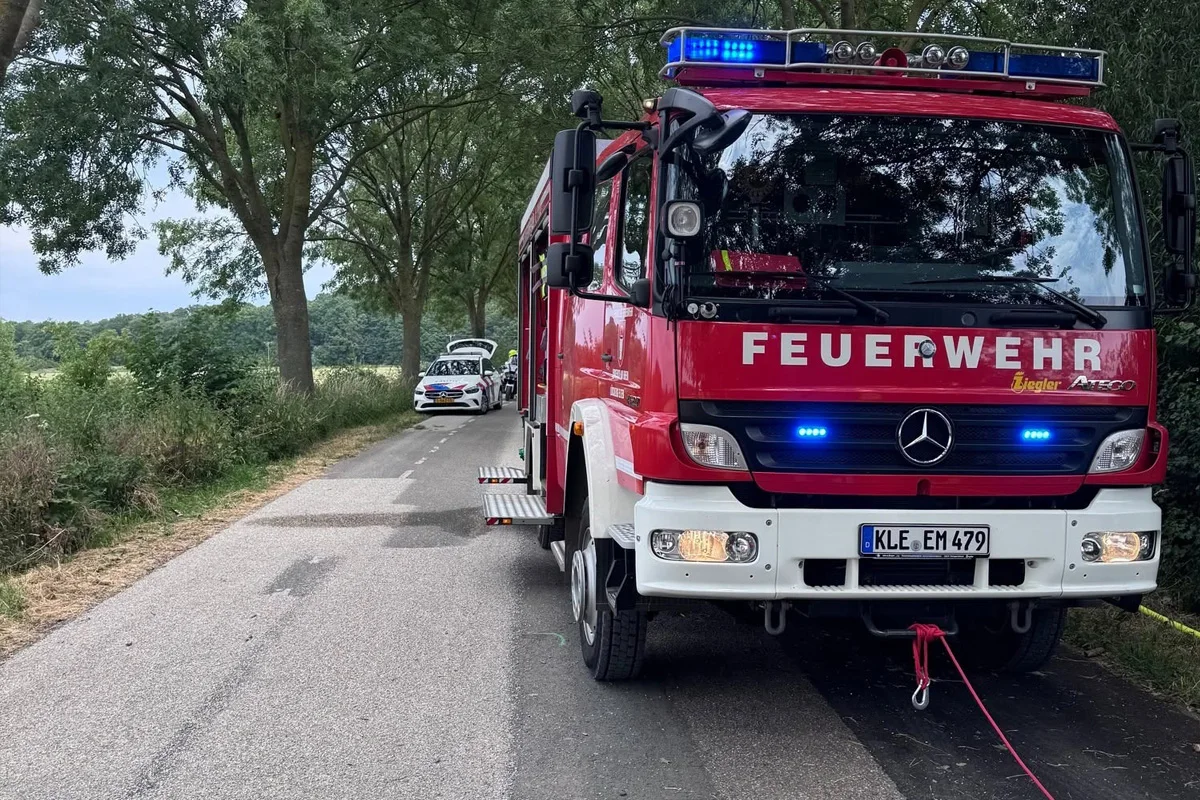 Ein rotes Feuerwehrauto steht am Straßenrand, dahinter ein Polizeiwagen. Die Szene ist von Bäumen gesäumt.