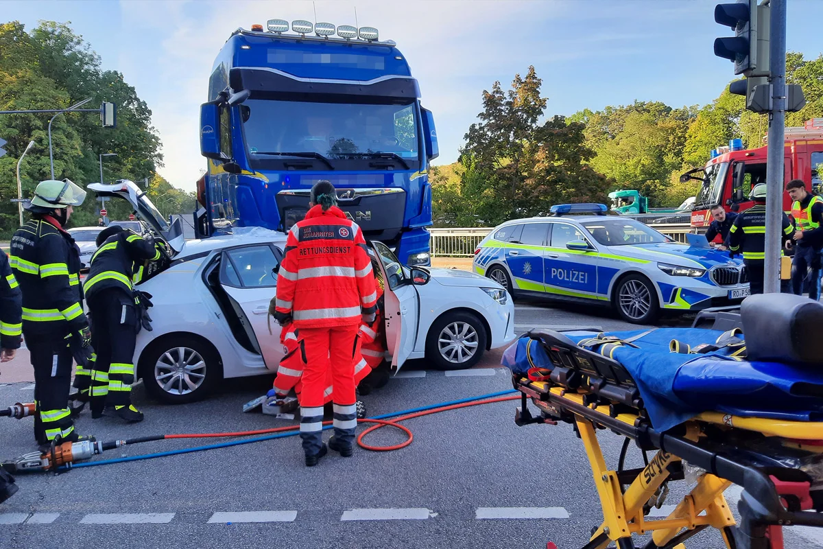 Ein blauer Lkw ist auf ein weißes Auto aufgefahren. Rettungskräfte arbeiten am Unfallort, ein Polizeiwagen steht nahebei.