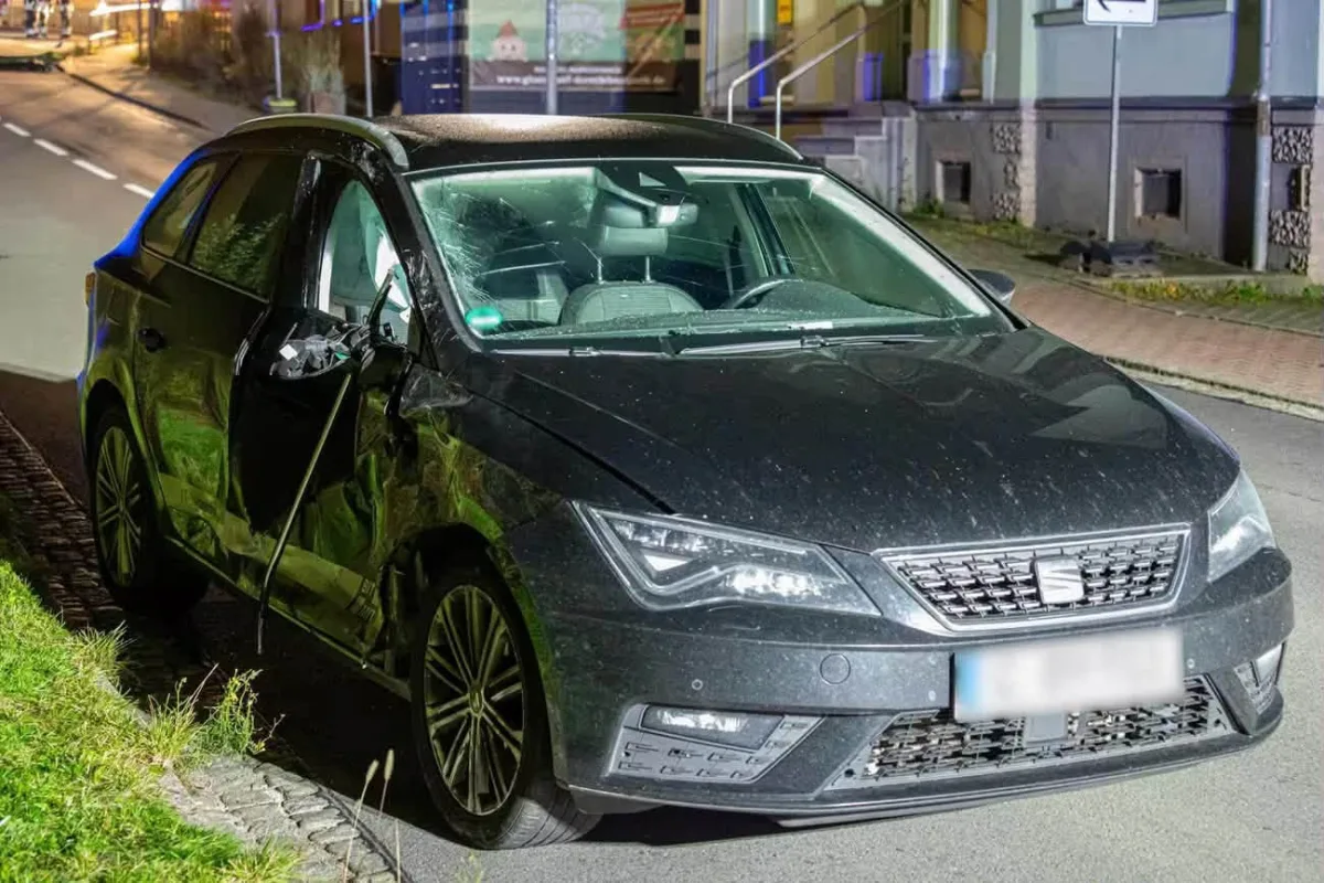 Ein schwarzer Pkw zeigt schwere Schäden auf der Fahrerseite. Es steht in einer städtischen Straße am Gehweg.
