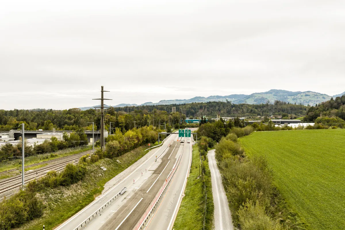 Das Bild zeigt einen freien Autobahnabschnitt, eingekreist von grünen Feldern und Bäumen, mit fernen Bergen am Horizont.