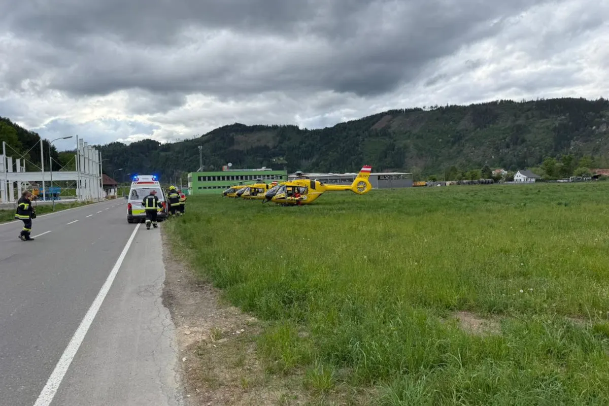 Mehrere gelbe Rettungshubschrauber stehen auf einem grünen Feld. Rettungskräfte sind bei einem weißen Fahrzeug auf einer angrenzenden Straße tätig.