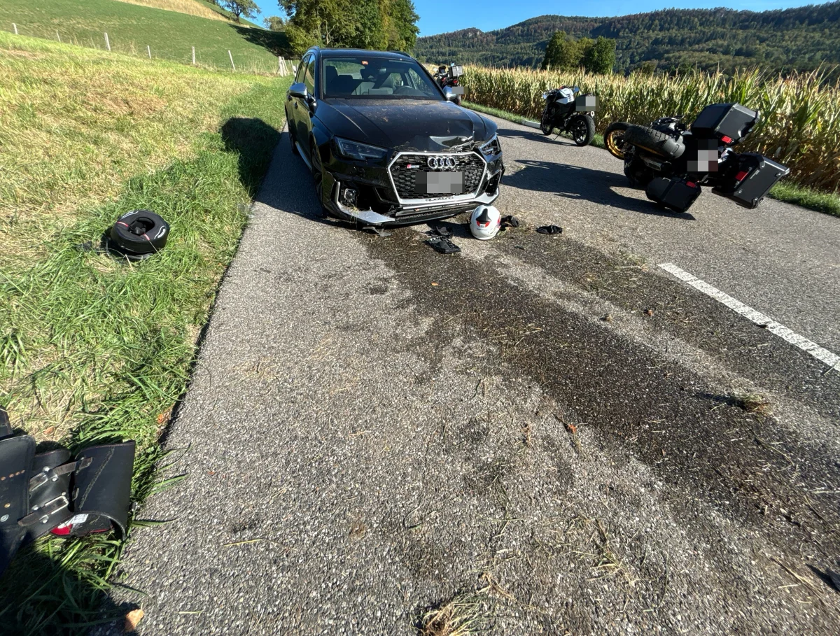 Ein schwarzer Audi steht beschädigt auf einer Landstraße neben einem umgestürzten Motorrad. Helme liegen verstreut auf der Fahrbahn.