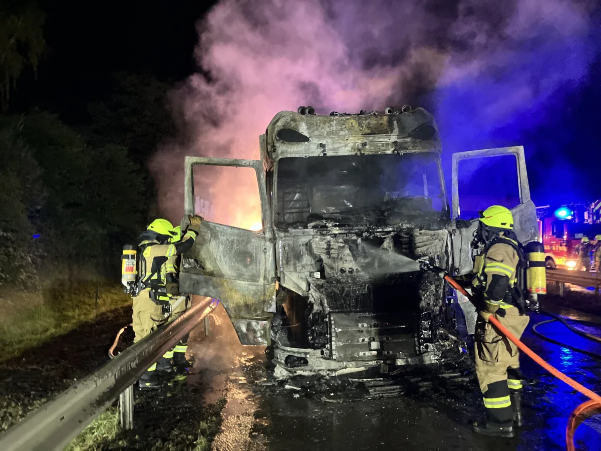 Zwei Feuerwehrleute in Schutzkleidung löschen bei Nacht mit Schläuchen einen ausgebrannten Lkw. Der Lkw steht auf einer Straße, umgeben von Rauch.