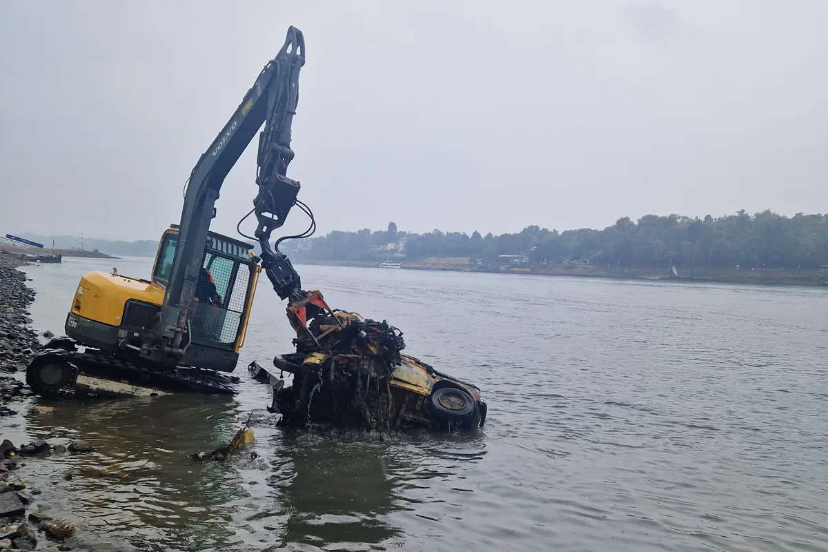 Ein gelber Bagger hebt ein stark verrostetes Auto aus einem Fluss, während er am Ufer steht. Der Himmel ist bewölkt.