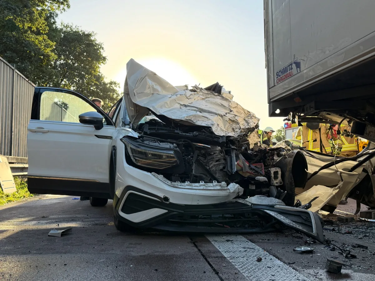 Ein weißes Auto mit schwer beschädigter Front ist frontal auf einen Lkw-Anhänger aufgefahren. Rettungskräfte sind im Hintergrund sichtbar.
