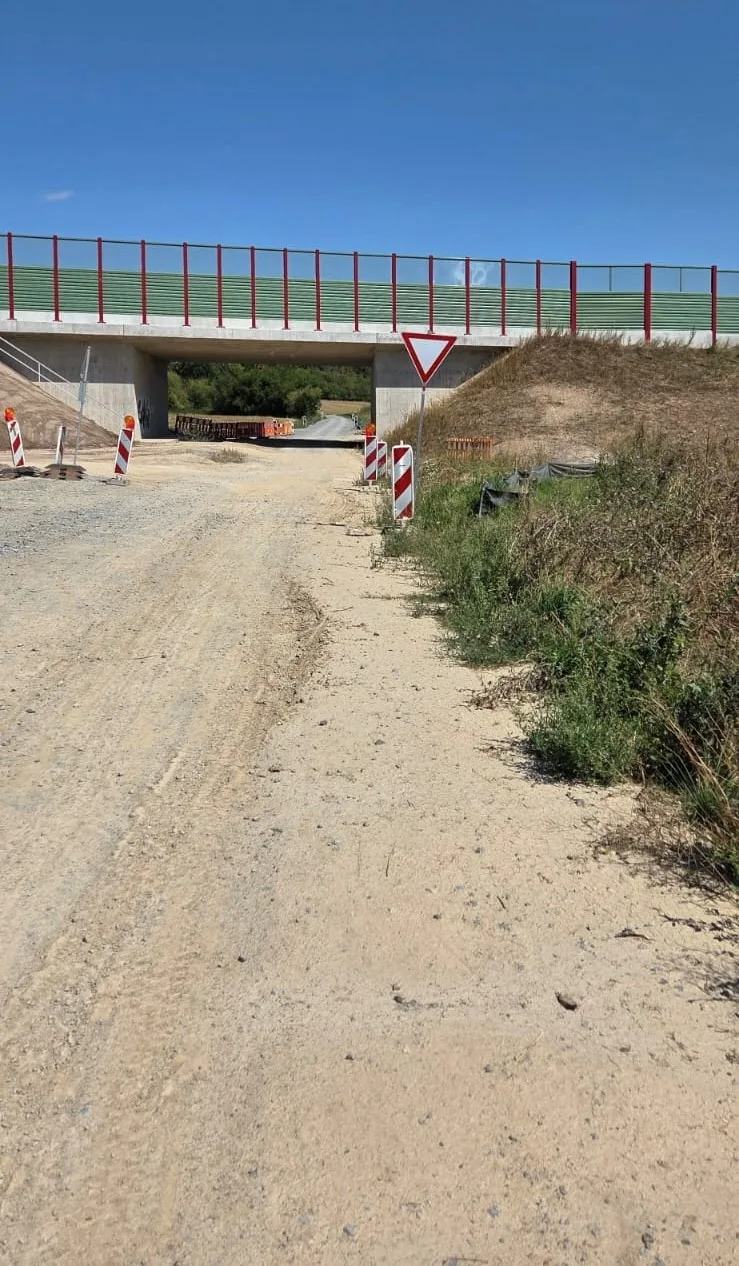 Eine unbefestigte Straße führt unter einer Brücke hindurch. Verkehrsschilder und Baustellenabsperrungen sind sichtbar.