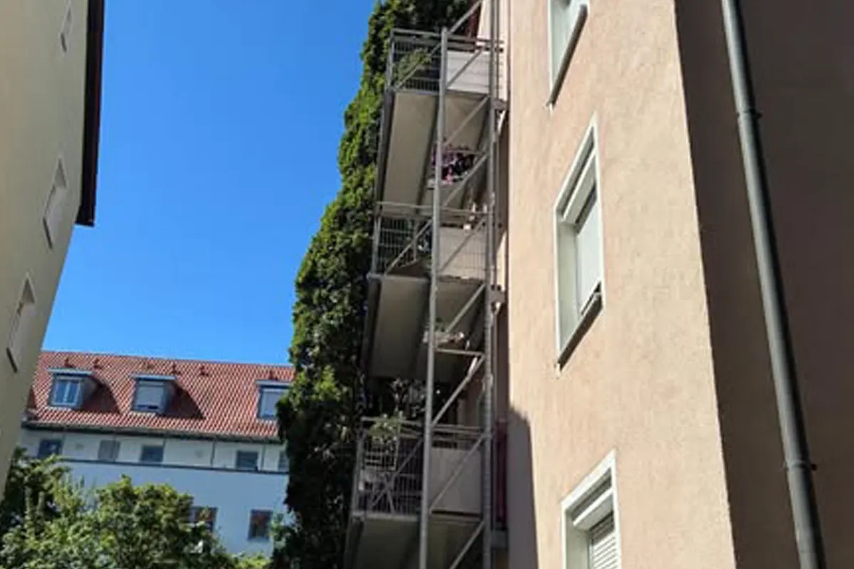 Mehrgeschossiges Wohnhaus mit Balkonen, umgeben von einer großen grünen Kletterpflanze, bei strahlend blauem Himmel.