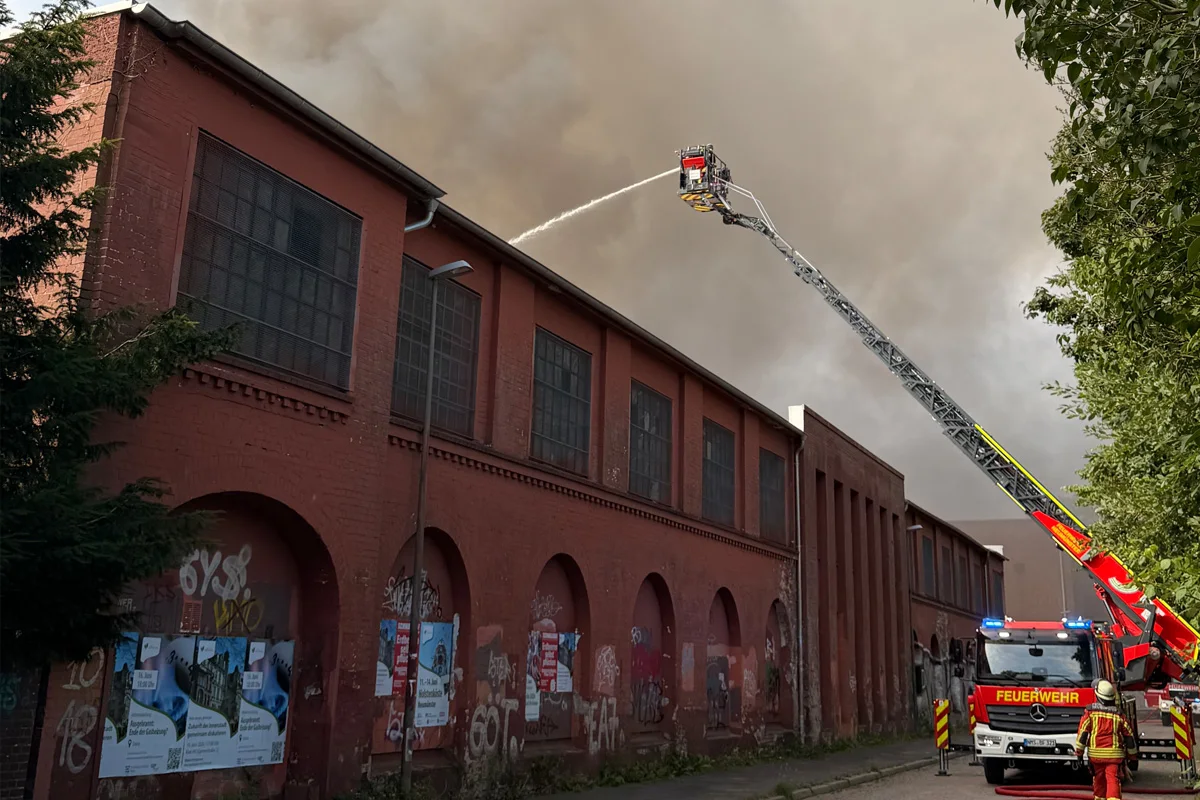 Ein Feuerwehrfahrzeug steht vor einer großen, dunklen Industriehalle. Ein Feuerwehrmann auf der Drehleiter löscht den Brand.