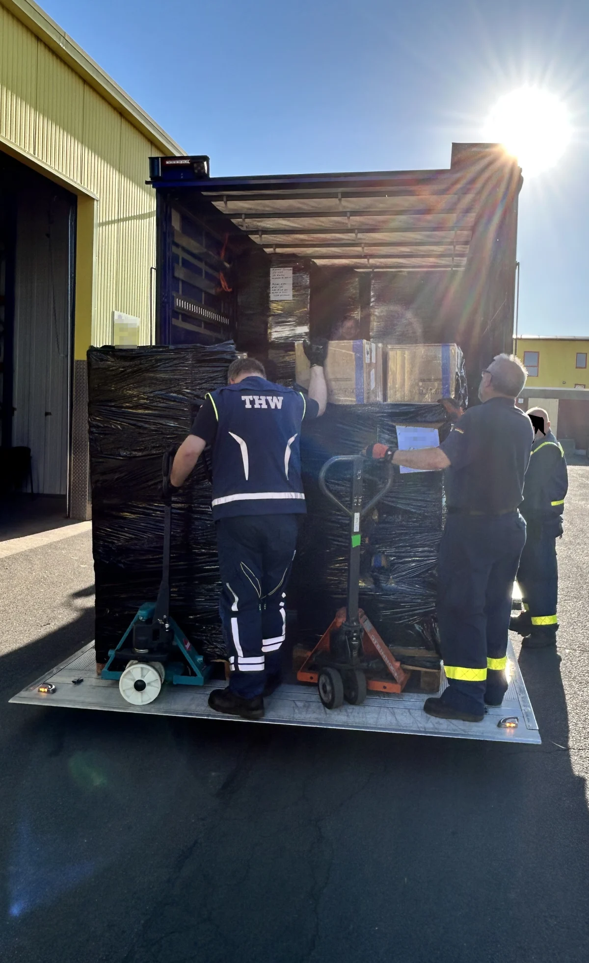 Drei Personen, erkennbar am THW-Aufdruck ihrer Uniformen, verladen mit Hubwagen große, in Plastik verpackte Paletten auf einen LKW. Die Sonne scheint hell auf die Szene.
