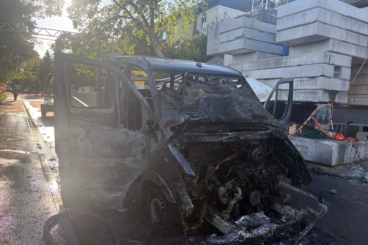 Ein stark beschädigter und ausgebrannter Van steht auf einer Straße. Im Hintergrund sind Baustellenmaterialien gestapelt.