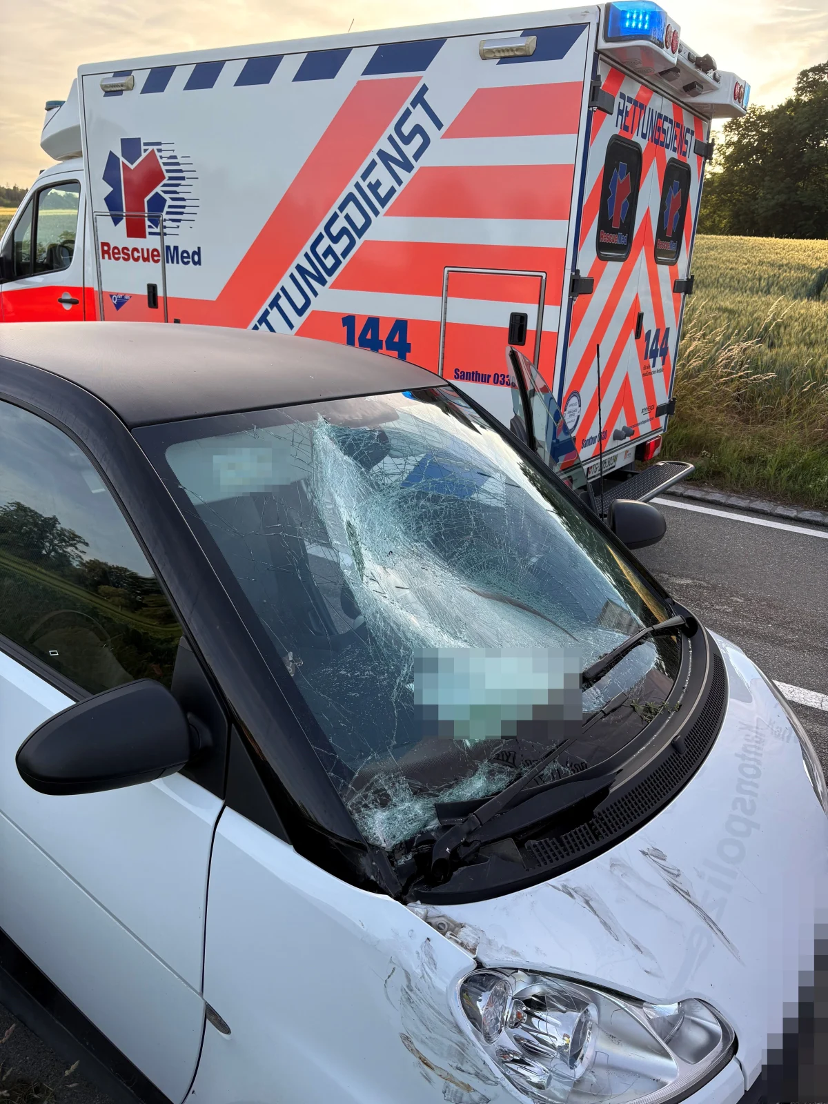 Ein weißer Smart mit beschädigter Windschutzscheibe steht neben einem Rettungswagen auf einer Landstraße. Im Hintergrund sind Felder zu sehen.