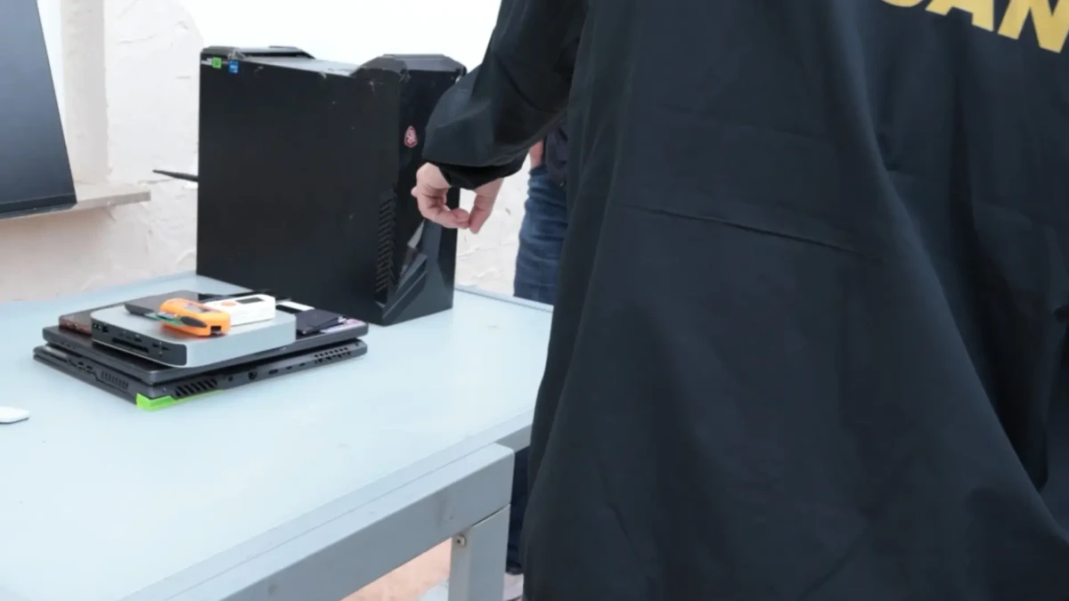 Auf einem Tisch liegen mehrere elektronische Geräte wie Computer und Laptops. Eine Person steht im Hintergrund, teilweise sichtbar.