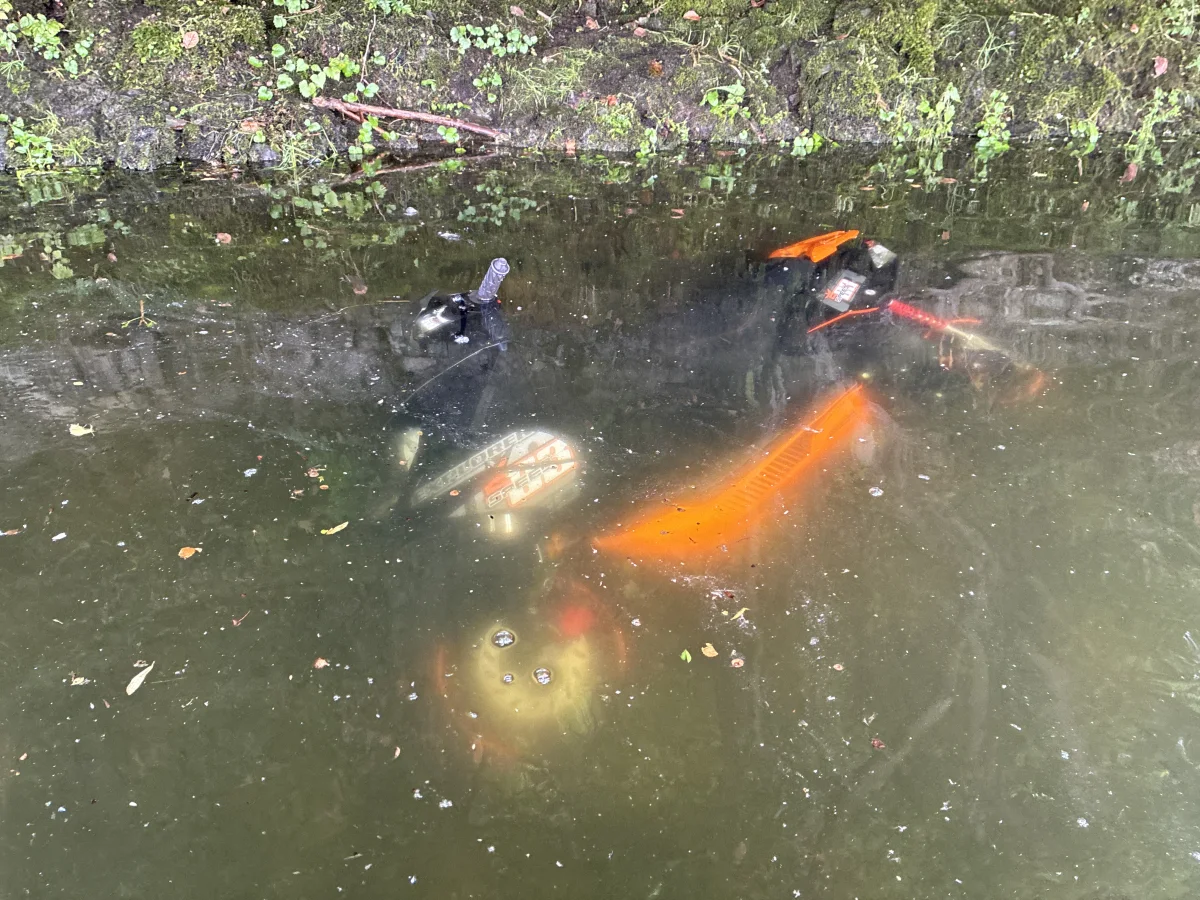 Ein orangefarbenes Motorrad liegt im Wasser eines Flusses. Teile des Motorrads sind nur teilweise sichtbar unter der Wasseroberfläche.