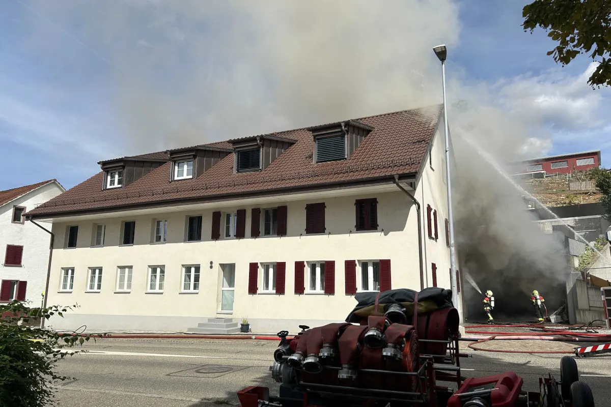 Dichter Rauch entweicht aus einem Gebäude, zwei Feuerwehrleute sind vor dem Haus im Einsatz mit Löschschläuchen.