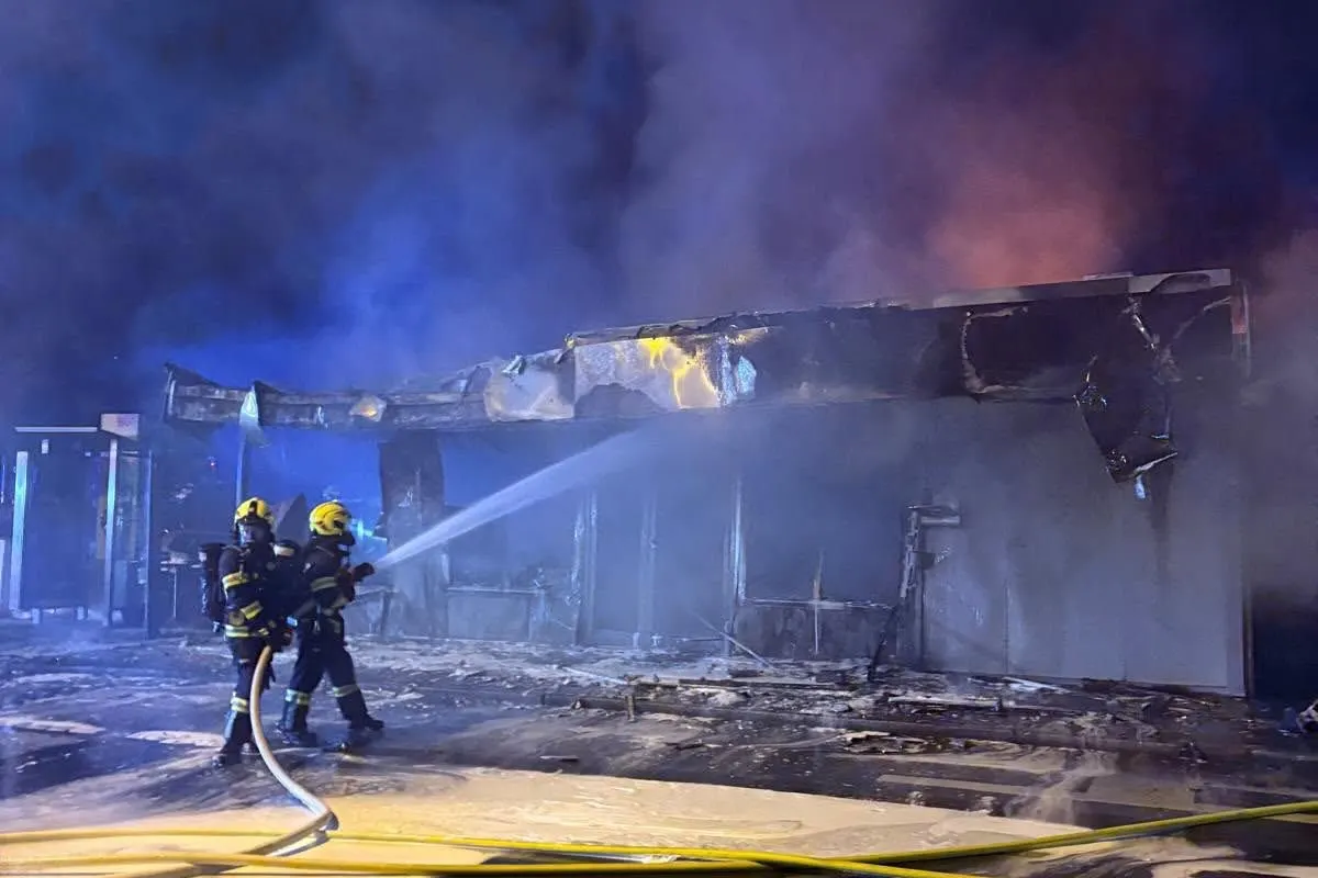 Zwei Feuerwehrleute in Schutzanzügen löschen bei Nacht ein brennendes Gebäude. Flammen und Rauch sind deutlich sichtbar.
