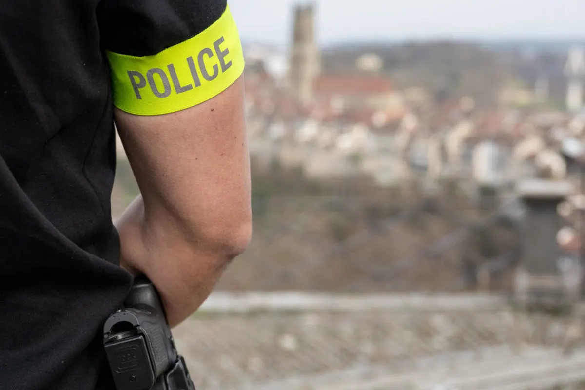 Ein Polizistenarm mit einer gelben Armbinde, auf der 'POLICE' steht, ist in der Bildmitte. Unscharfe Stadtansicht im Hintergrund.