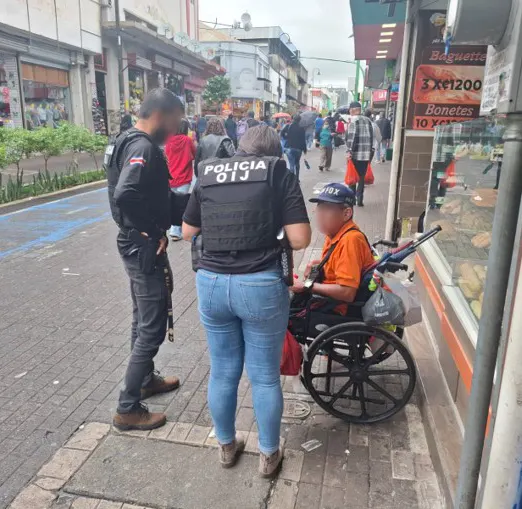 Zwei Polizisten unterhalten sich mit einem Mann im Rollstuhl in einer belebten Einkaufsstraße mit vielen Passanten.