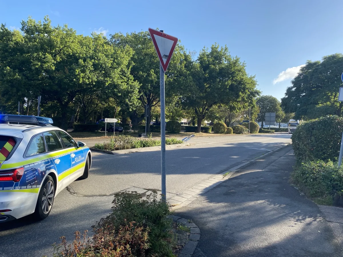 Ein deutsches Polizeifahrzeug steht nahe einer Kreuzung. Die Szenerie ist sonnig, mit grünem Laub im Hintergrund.