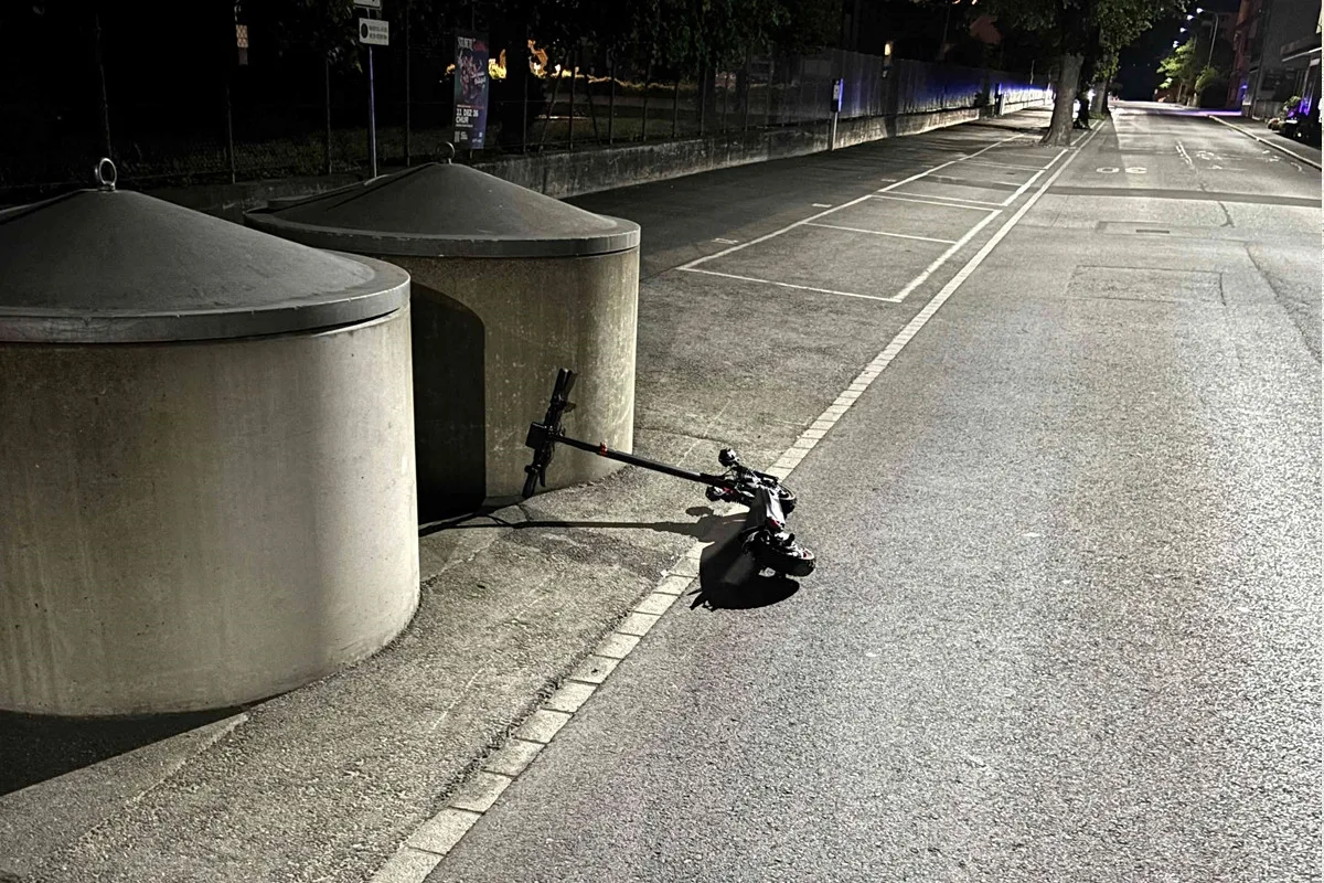 Ein umgefallener E-Scooter liegt neben zwei großen Betonbehältern auf einem Gehweg. Die Szene spielt sich bei Nacht in einer ruhigen Straße ab.