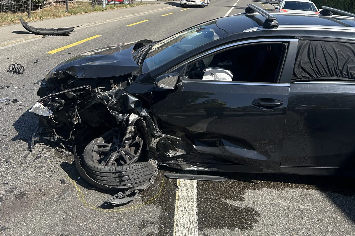 Ein schwarzes Auto zeigt schweren Frontschaden nach einem Unfall auf der Straße. Der Airbag ist ausgelöst, und es liegen Trümmerteile auf der Fahrbahn.