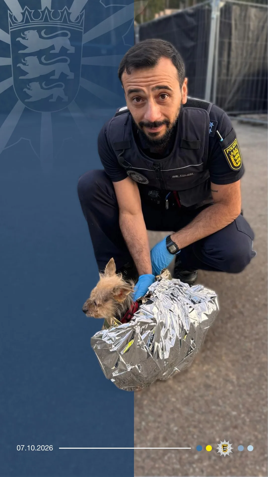 Ein Polizist kniet auf dem Boden und hält einen kleinen Hund, der in eine silberne Rettungsdecke eingewickelt ist, wahrscheinlich nach einer Rettungsaktion.