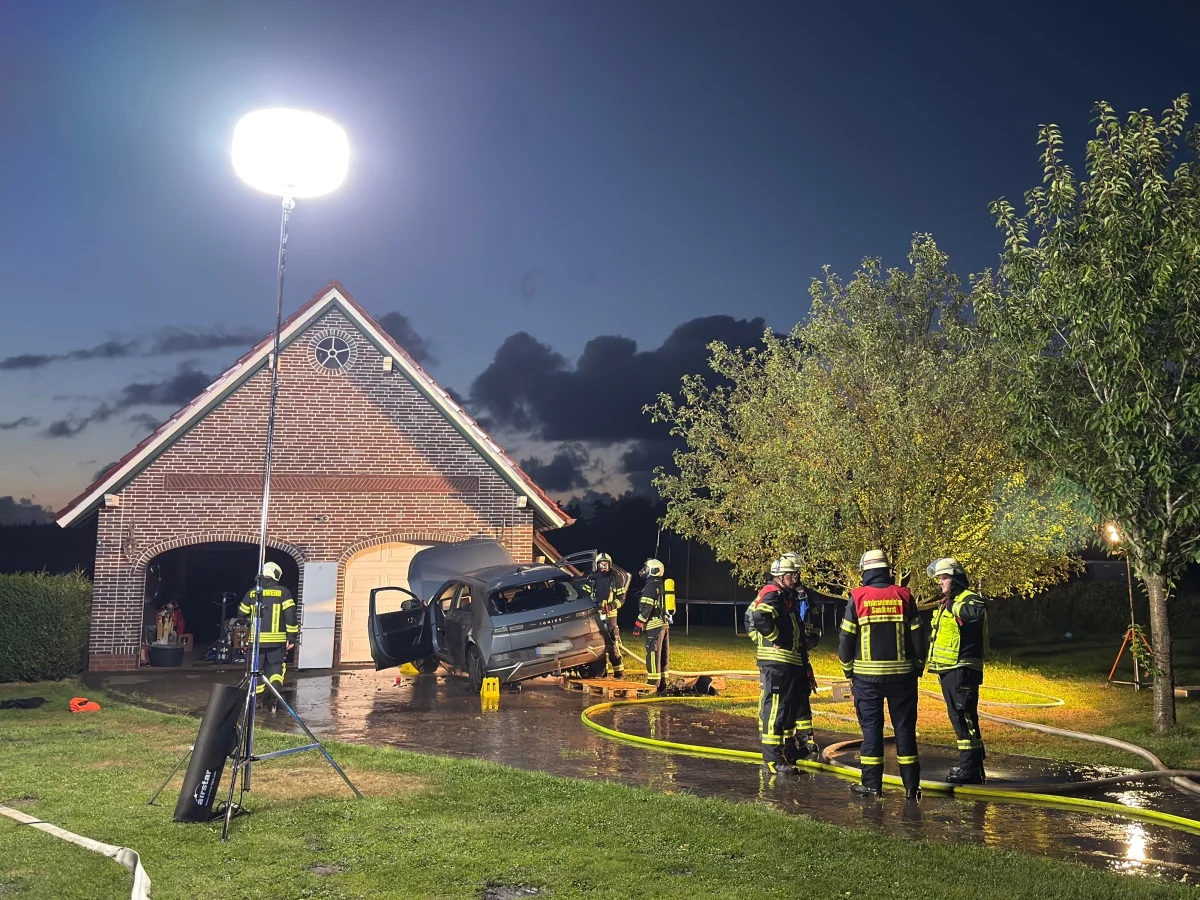 Ein Auto mit offener Heckklappe steht beschädigt vor einer Garage. Mehrere Feuerwehrleute stehen in der Nähe unter einer großen Einsatzleuchte.
